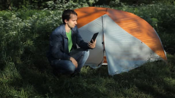 Mężczyzna siedzi w pobliżu namiotu i robi rozmowę wideo na tablecie. Rano w lesie — Wideo stockowe