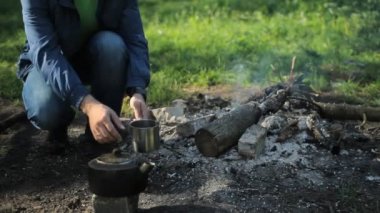 Adam bir demlikten metal bir bardağa çay döker. Yangının yanında yanıklar, sabahın erken saatlerinde ormanda