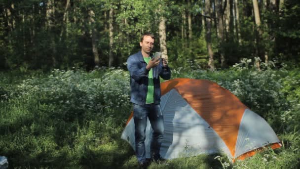 Bir adam telefonda görüntülü arama yapıyor. Yakındaki ormanda bir çadır duruyor, sabahın erken saatlerinde — Stok video