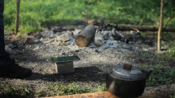 Tablette auf einen Stein in der Nähe des rauchenden Feuers gelegt. Im Vordergrund ein Wasserkocher. frühmorgens im Wald — Stockvideo