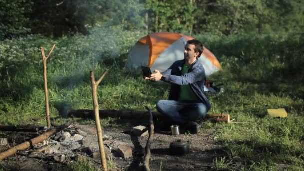 Мужчина фотографирует пожар на планшете. На заднем плане палатка в лесу рано утром — стоковое видео