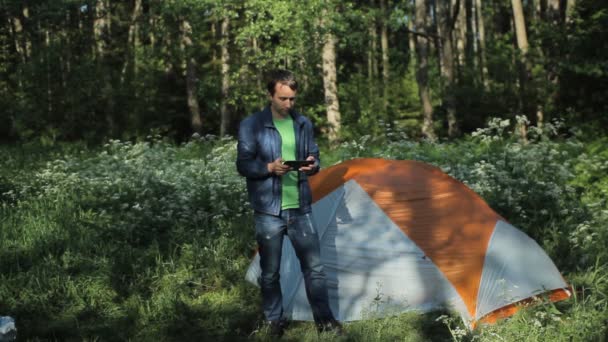 En man som leker på en surfplatta bredvid tältet i skogen, tidigt på morgonen — Stockvideo