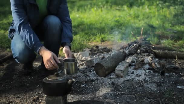 Man pours tea from a pot into a metal cup. Near the fire burns, the early morning in the forest — Stock Video