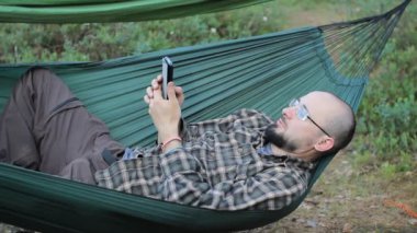 Bir hamakta telefonda çağrı video biri. Adam bir sakal ve gözlük ile ormanda