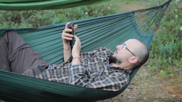 Muž v houpací síti dotykový telefon. Muž v lese s plnovousem a brýlemi — Stock video