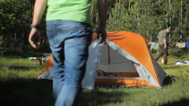 Człowiek w obozie przychodzi do namiotu i zamiar odpocząć. Na tle lasu ludzie chodzą na słoneczny dzień — Wideo stockowe