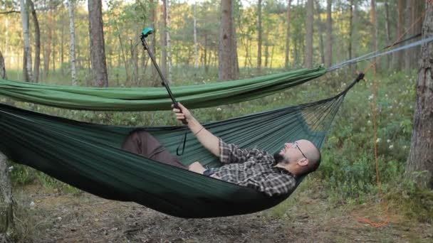 Een man in een hangmat op selfie stick met actiecamera video te maken. Man in het bos met een baard en glazen — Stockvideo