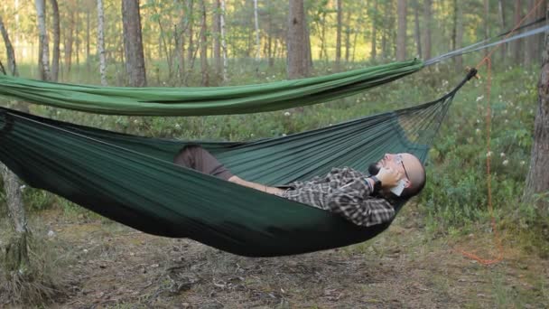 Seorang pria di tempat tidur gantung menelepon melalui telepon. Pria di hutan dengan jenggot dan kacamata — Stok Video
