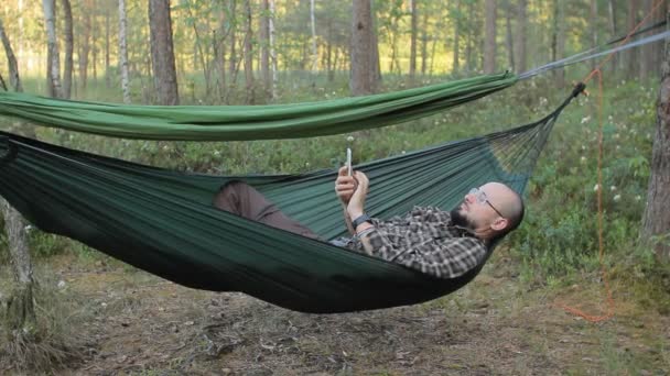 Un hombre en un teléfono táctil hamaca. Hombre en el bosque con barba y gafas — Vídeos de Stock