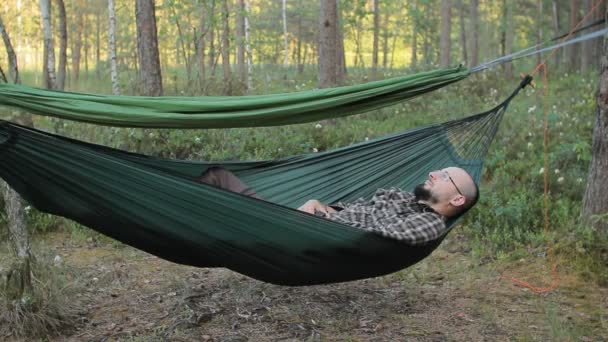 Een man in een hangmat touch smartwatch. Man in het bos met een baard en glazen — Stockvideo