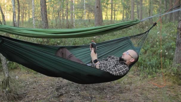 Een man in een hangmat touch tablet. Man in het bos met een baard en glazen — Stockvideo