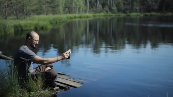 Un homme est assis sur une jetée du lac et fait un portrait selfie au téléphone avec un bâton de selfie. Le beau lac bleu et la forêt sur le fond — Video