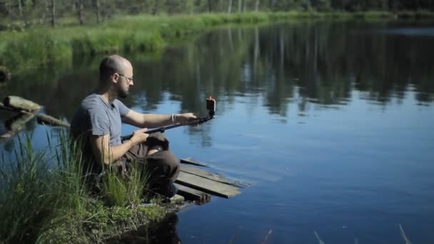 Un hombre se sienta en un muelle del lago y hace un retrato de selfie por teléfono con un palo de selfie. El hermoso lago azul y el bosque en el fondo — Vídeos de Stock
