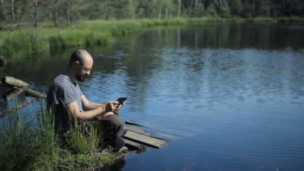 En man sitter på en brygga i sjön och spela på tablett i spelet. Den vackra blå sjön och skogen på bakgrunden — Stockvideo
