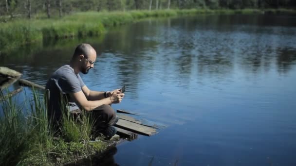 Um homem senta-se num cais do lago e toca no tablet. O belo lago azul e floresta no fundo — Vídeo de Stock