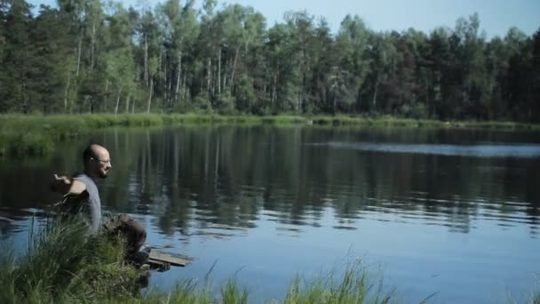 Um homem senta-se em um cais do lago e a beleza das maravilhas. O belo lago azul e floresta no fundo — Vídeo de Stock