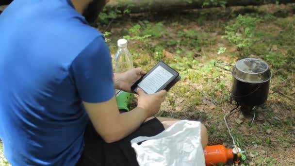 Man läser en e-bok i skogen. Nära turist potten på en gasbrännare — Stockvideo