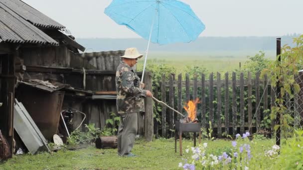 Man met paraplu in de buurt van het vuur voor barbecue. De regen valt, een oud gebouw met een hek op de achtergrond — Stockvideo