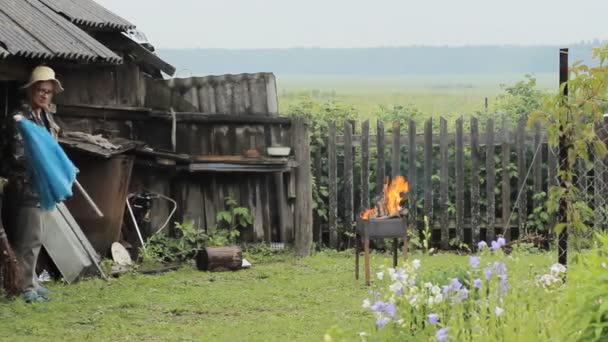 Чоловік з парасолькою біля багаття для барбекю. Дощ падає, стара будівля з парканом на задньому плані — стокове відео