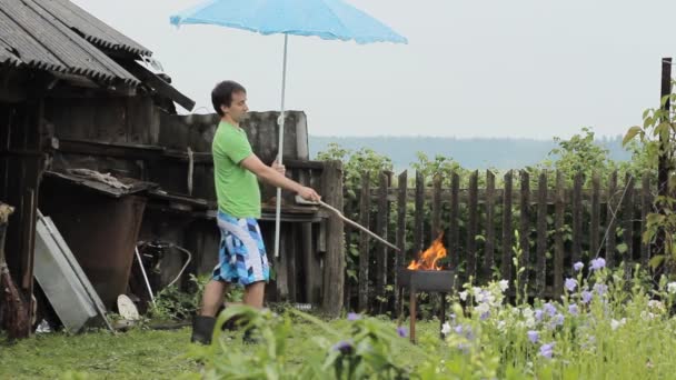 Man with umbrella near the fire for barbecue. The rain is falling, an old building with a fence in the background — Stock Video