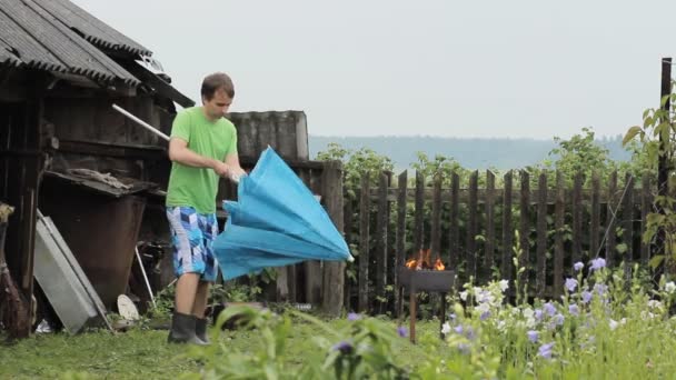 Чоловік з парасолькою біля багаття для барбекю. Дощ падає, стара будівля з парканом на задньому плані — стокове відео