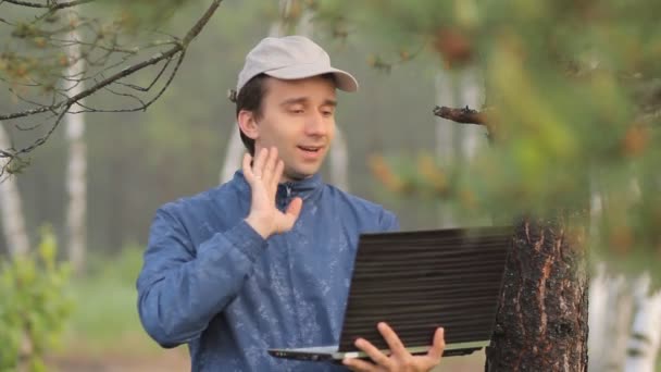 Ein Mann telefoniert mit einem Laptop in der Nähe eines Baumes im Wald. frühmorgens — Stockvideo