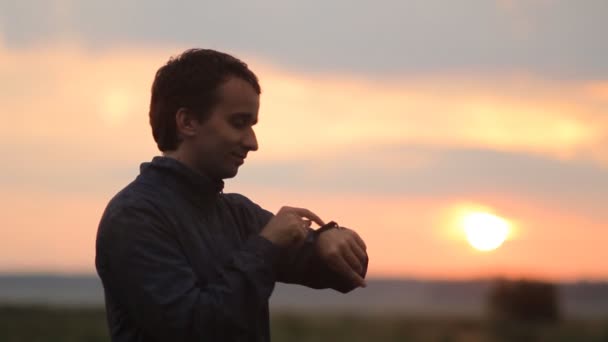 El hombre toca el reloj inteligente. Hermosa puesta de sol en el fondo — Vídeos de Stock