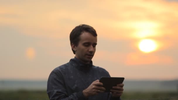Un hombre jugando en una tablet en el juego. Hermosa puesta de sol en el fondo — Vídeos de Stock