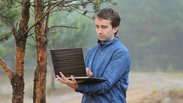 En man som arbetar på en bärbar dator nära ett träd i skogen. Tidigt på morgonen — Stockvideo