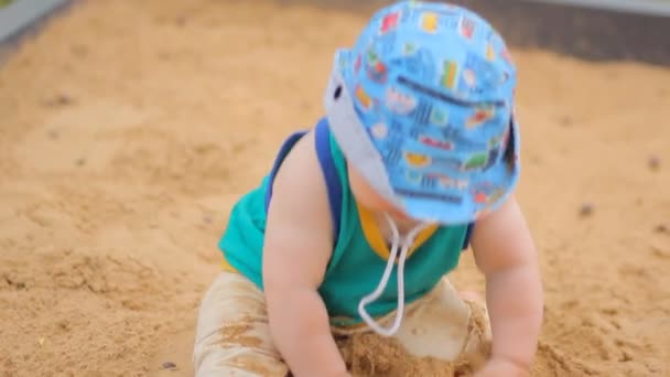 Rapaz a cavar na caixa de areia. Sorri e toca a areia — Vídeo de Stock