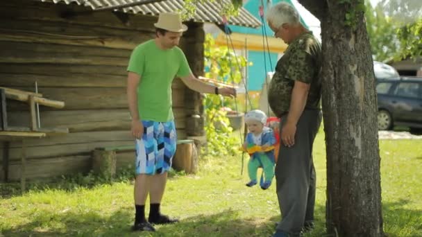 年配の男性と父親は、ブランコに乗って美しい赤ちゃんをロールバックします。彼の家の庭で孫と祖父 — ストック動画