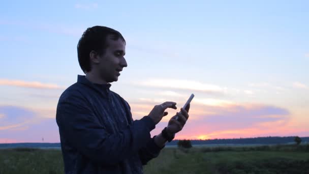 Молодий чоловік тримає мобільник на заході сонця. Він пише смс — стокове відео