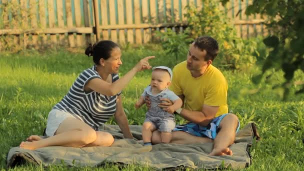 Счастливая семья из трех человек лежит в траве в летнем парке. Они играют и улыбаются. — стоковое видео