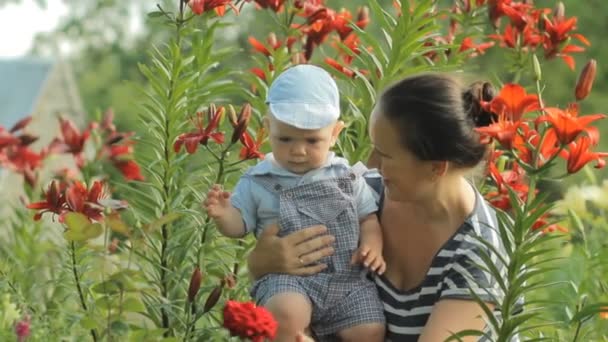 Joven madre con su hermoso niño tocando flores cerca de su casa. El hijo mira las plantas con atención — Vídeos de Stock