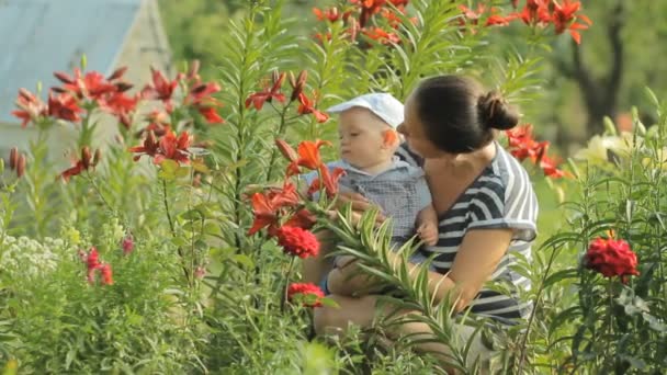 Joven madre con su hermoso niño tocando flores cerca de su casa. El hijo mira las plantas con atención — Vídeos de Stock