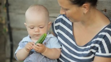 Güzel bebek erkek bebek anne ellerde oturur ve salatalık yemek. Eski ahşap bir evin yanında