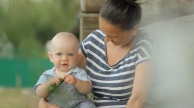 Güzel bebek erkek bebek anne ellerde oturur ve salatalık yemek. Eski ahşap bir evin yanında