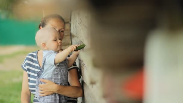 Il bel bambino si siede a mamma su mani e mangia il cetriolo. Vicino a una vecchia casa di legno — Video Stock