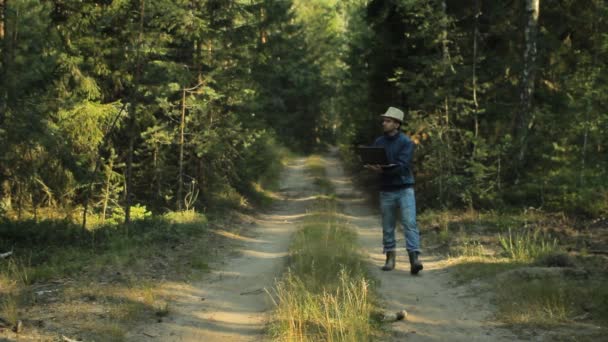 En man går genom skogen med en bärbar dator och ser kartan han väljer rätt riktning — Stockvideo