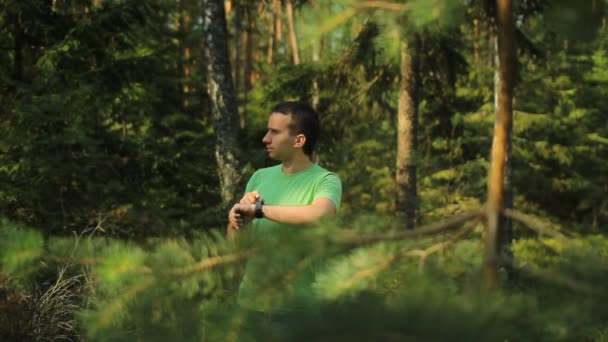 Un hombre camina por el bosque con un reloj inteligente y mirando la aplicación de navegación. Él elige la dirección correcta — Vídeos de Stock