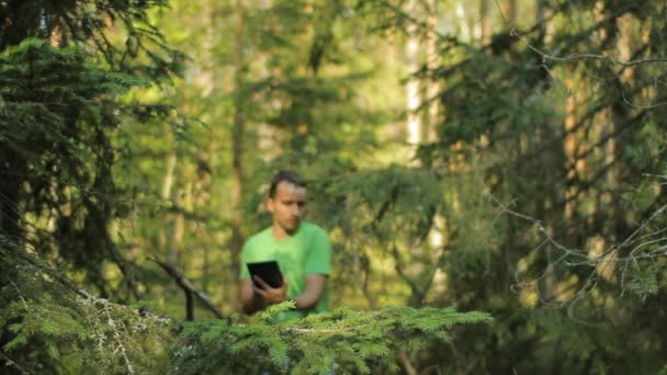 Seorang pria berjalan melalui hutan dengan tablet PC dan melihat aplikasi navigasi. Dia memilih arah yang benar — Stok Video