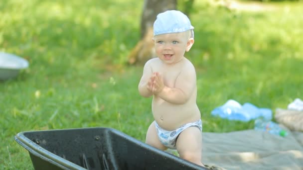 Petit garçon assis près du bassin dans un jardin verdoyant. Beau bébé souriant, frappe l'eau monte et se couche — Video