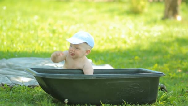 Малыш сидит возле бассейна в зеленом саду. Младенец улыбается, ударяется в воду и садится — стоковое видео