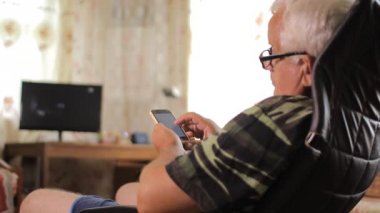 Bir telefonla ev Tv. Man yazarak sms ve izlerken fotoğraf yakınındaki bir deri sandalyede oturan üst düzey adam