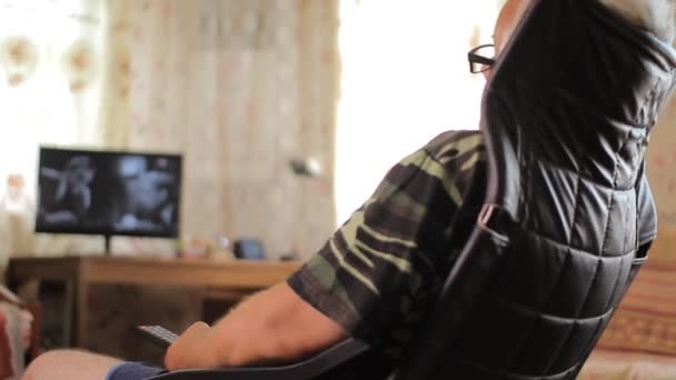 Homme âgé assis dans une chaise en cuir et change de chaînes TV télécommande à la maison — Video