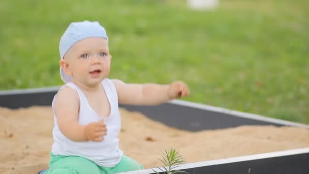 Schattige babyjongen spelen met zand in een zandbak. Zomer park en groen gras op de achtergrond — Stockvideo
