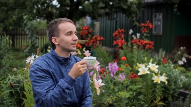 Hombre adulto bebe café de la mañana en el jardín, día de verano. Sobre el fondo de flores blancas, rojas, púrpuras — Vídeos de Stock