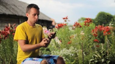 Genç adam oyun bahçesinde çiçekler yakınındaki için tablet kullanın. Beyaz, kırmızı ve mor çiçekler içinde belgili tanımlık geçmiş