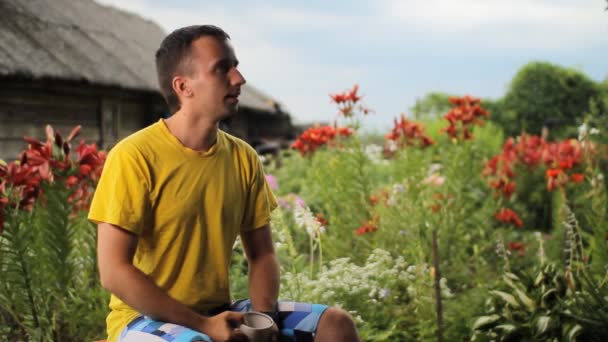 Vuxen man dricker morgonkaffe i trädgård, sommardag. Mot bakgrund av vitt, rött, lila blommor — Stockvideo