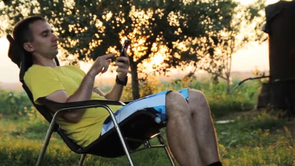 Jonge man, zittend in een stoel met een telefoon in een tuin. Tijdens pauzes bomen oranje zon bij zonsondergang — Stockvideo